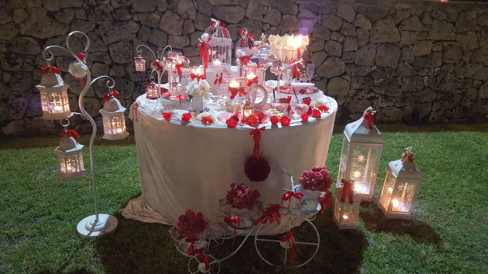 Tavolo della torta decorato con fiori rossi, candele e lanterne su un prato addossato a un muro di pietra.