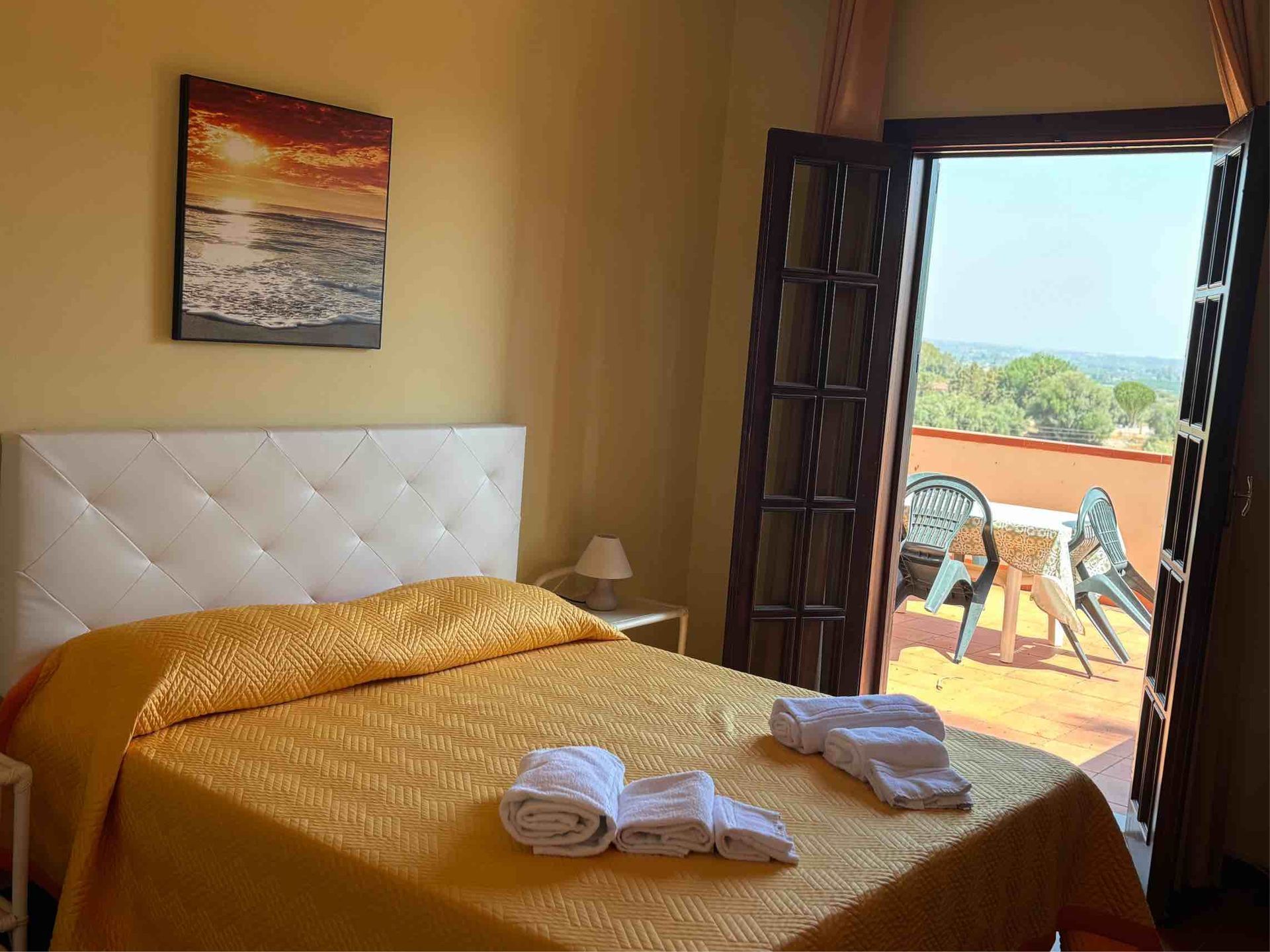 Camera da letto con letto, asciugamani piegati e porte aperte che conducono a un balcone soleggiato con vista mare.