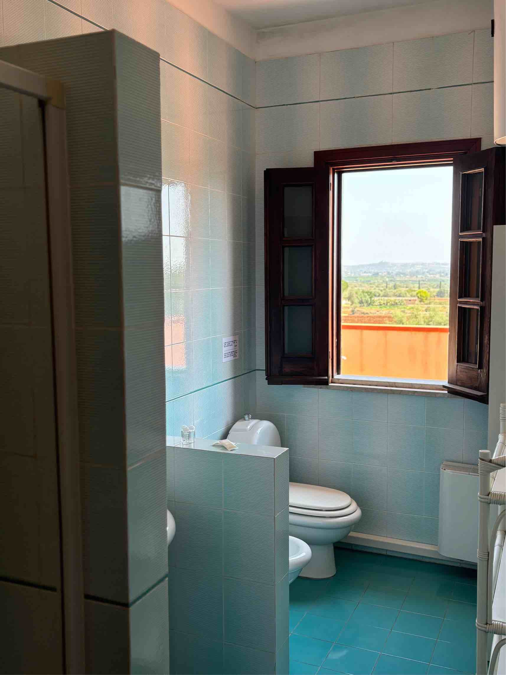 Piccolo bagno piastrellato con WC, finestra aperta e pavimento turchese, con vista sull'esterno.