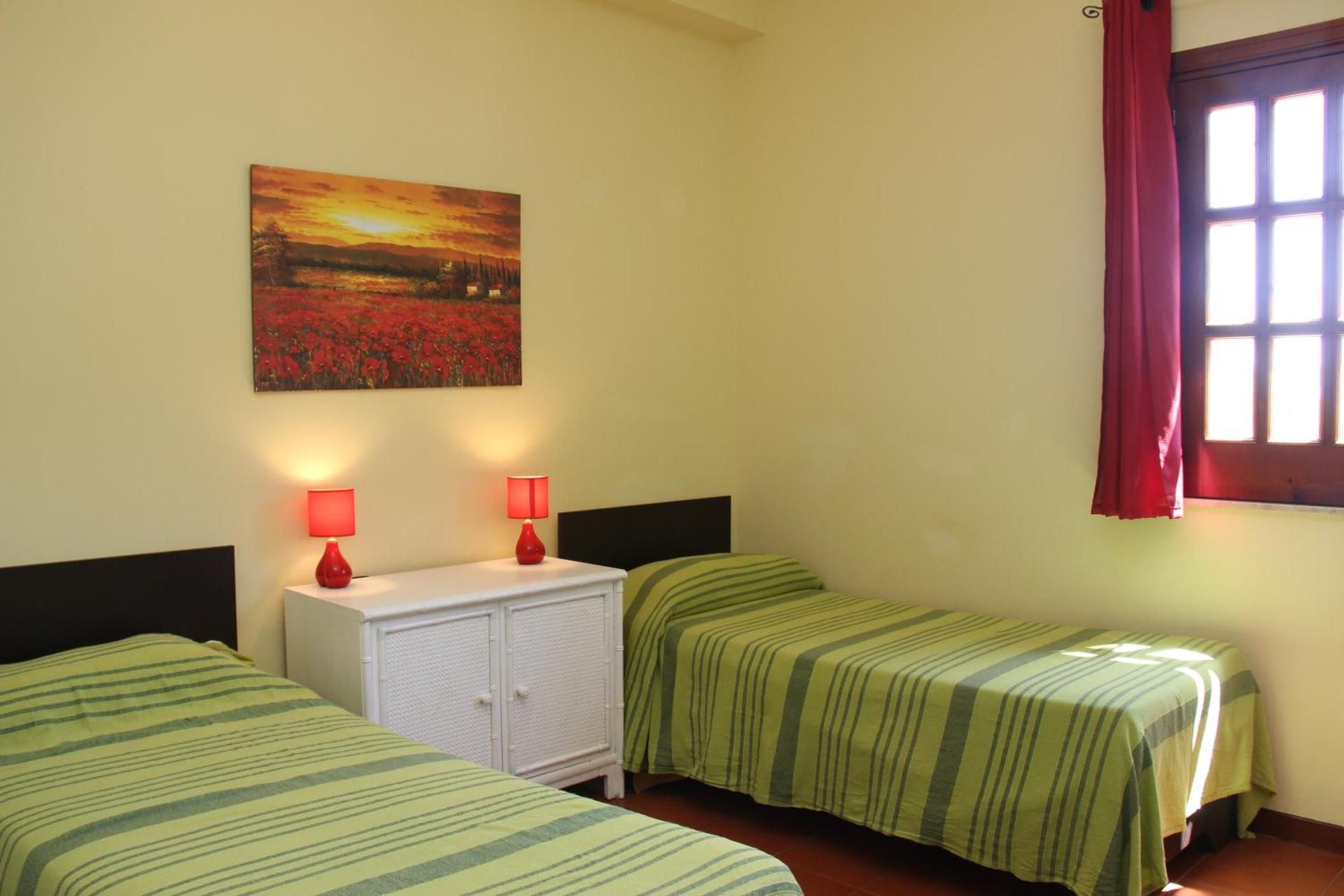 Camera da letto con due letti singoli, comodino bianco, lampade rosse e una finestra con tende rosa.