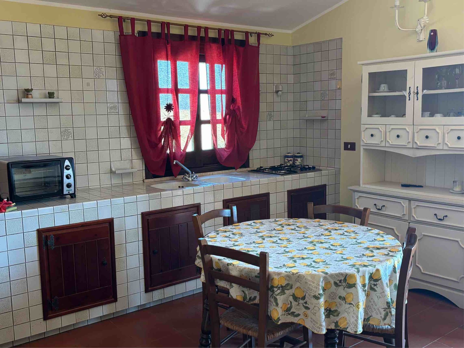 Piccola cucina con pareti rivestite di piastrelle a scacchi, tende rosse, un tavolo rotondo e mobili bianchi.