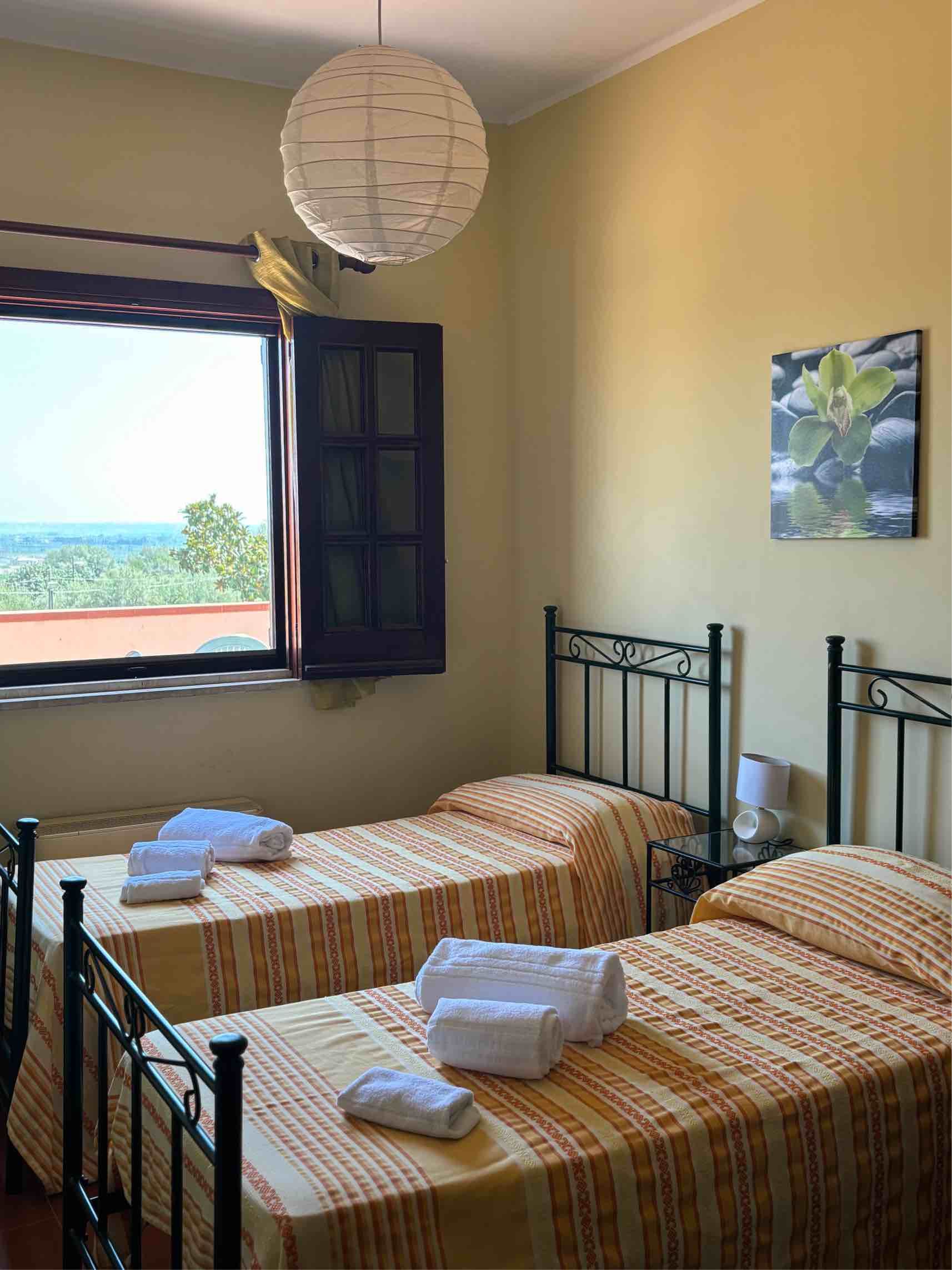 Piccola camera da letto con due letti singoli, copriletti a fantasia, una finestra con vista e una lampada a sospensione.