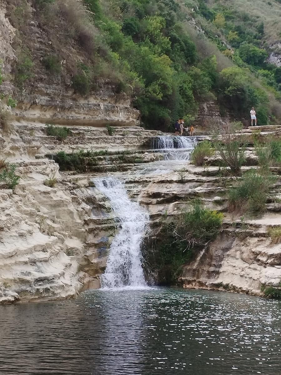 Una piccola cascata scende a cascata su strati di roccia formando una pozza, con arbusti verdi lungo il pendio.