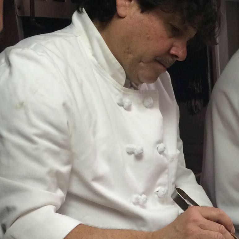 Chef in a white jacket writing with a pen, focused in a kitchen setting.
