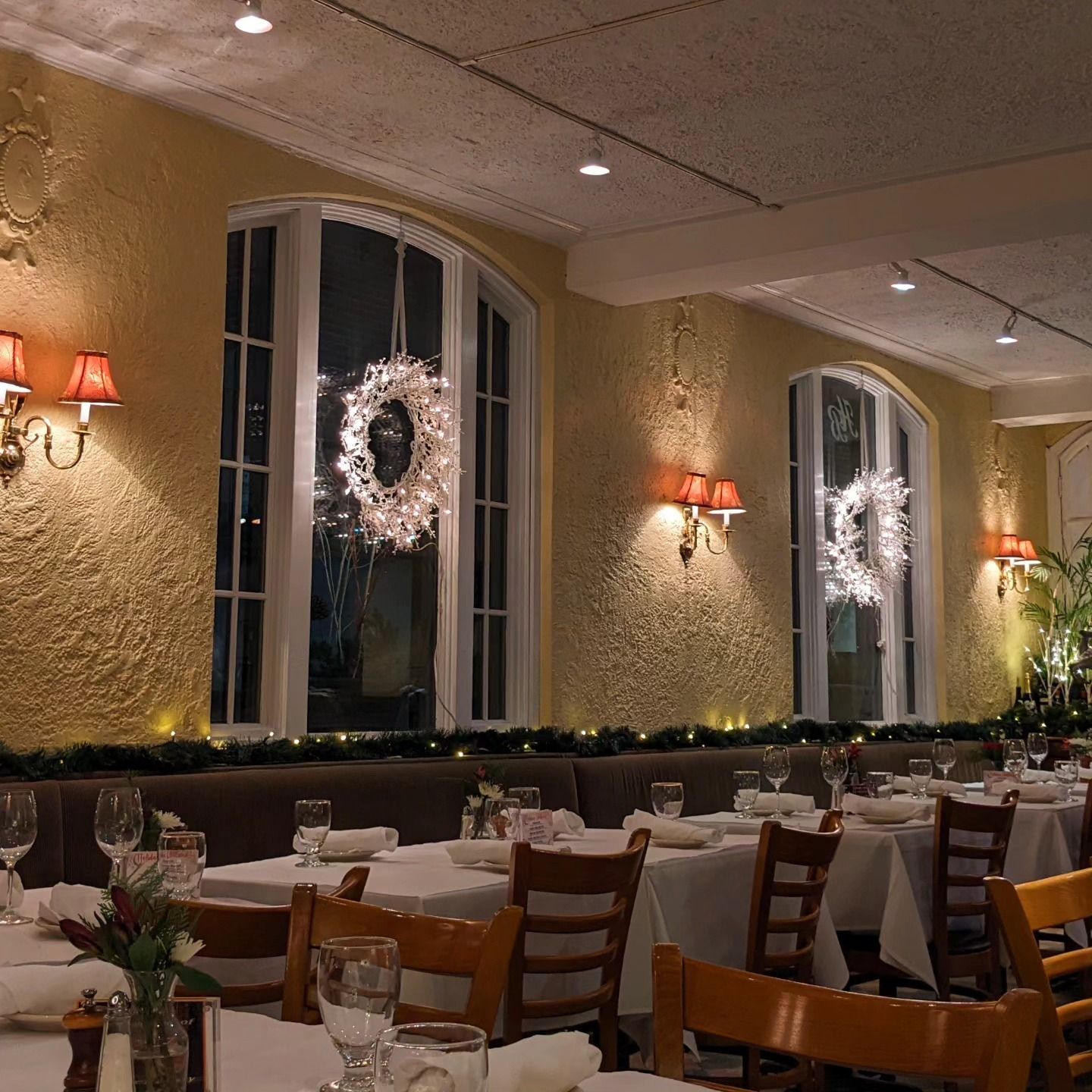 Elegant banquet hall with set tables, chandeliers, and holiday wreaths on tall windows