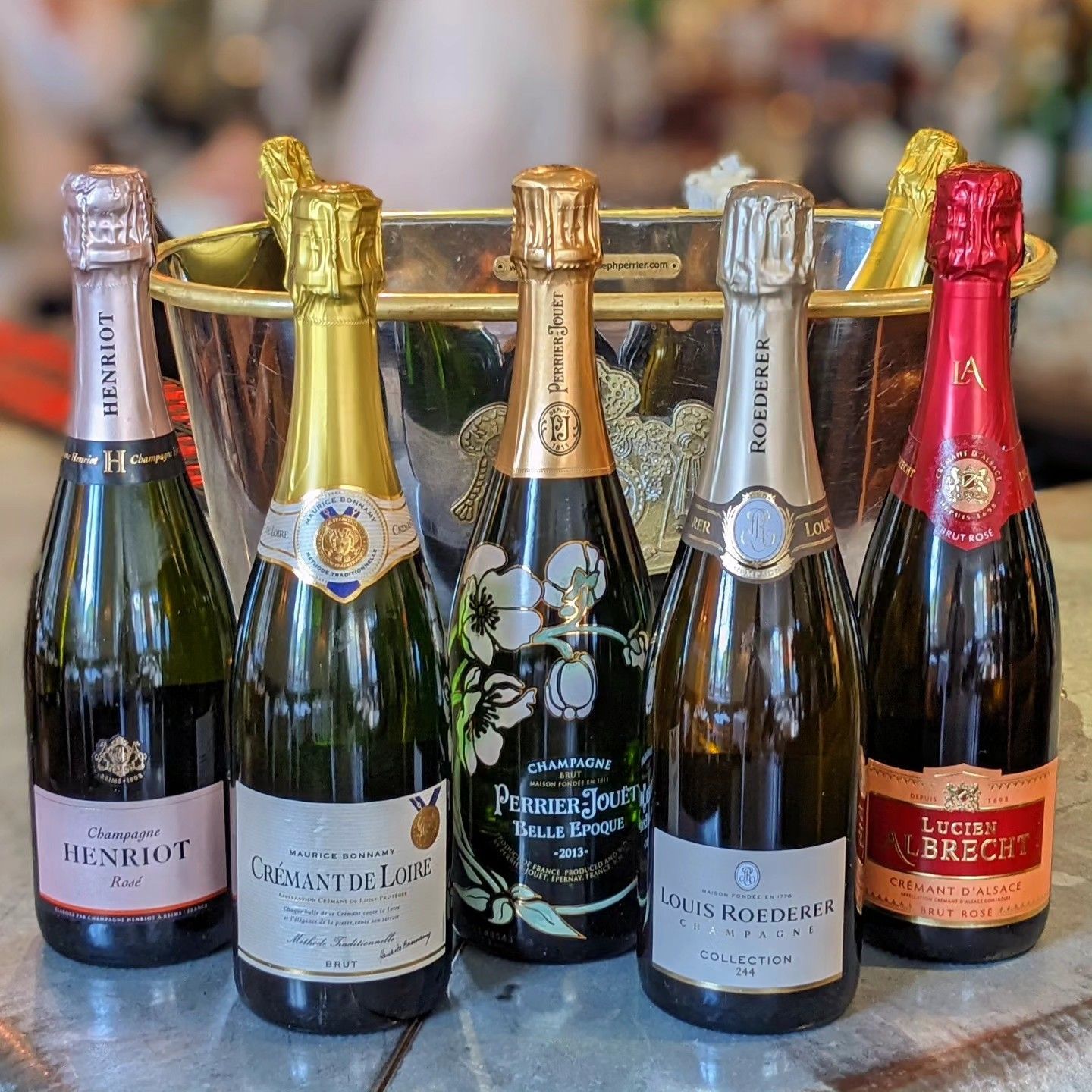 Five champagne bottles with gold and silver foil, lined up on a table against a blurred background.