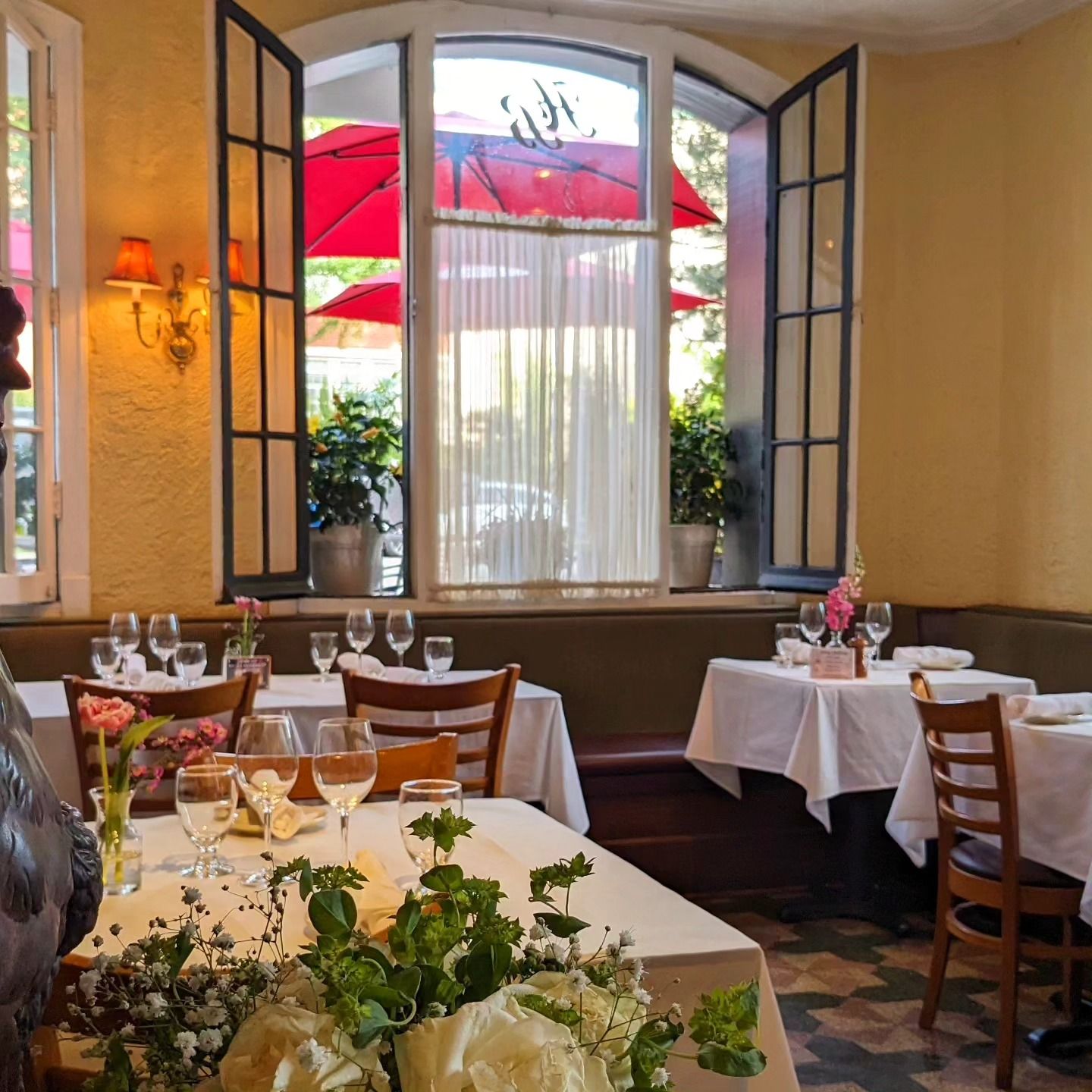 Elegant restaurant dining room with white tablecloths, red umbrellas outside, and sunlight through tall windows