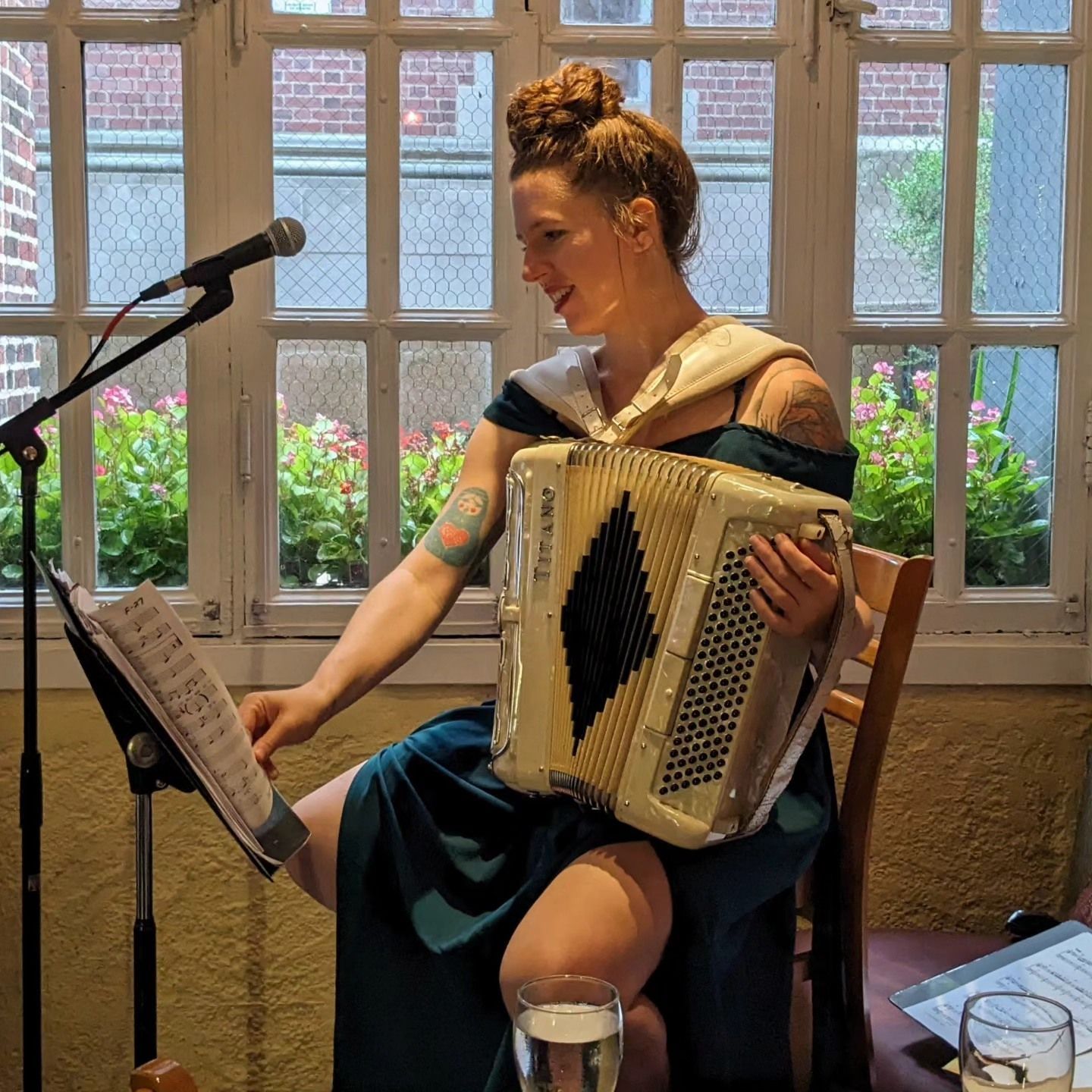 Woman playing accordion beside a microphone in a cozy indoor venue with large windows