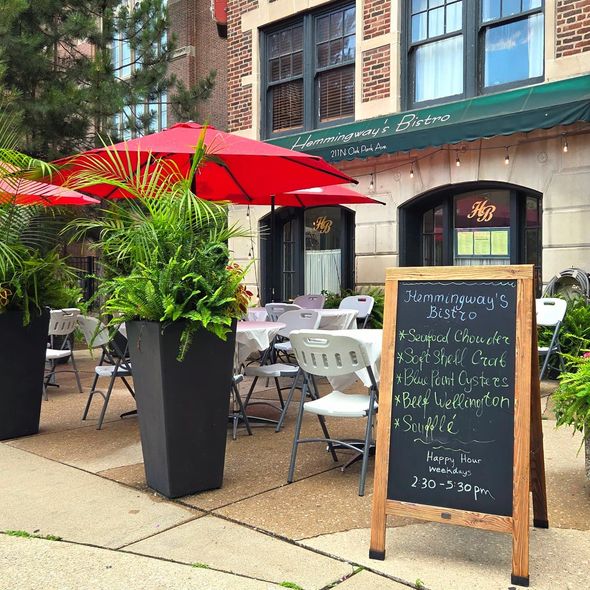 Outdoor café patio with red umbrellas, black planters, tables, and a chalkboard menu on a sidewalk