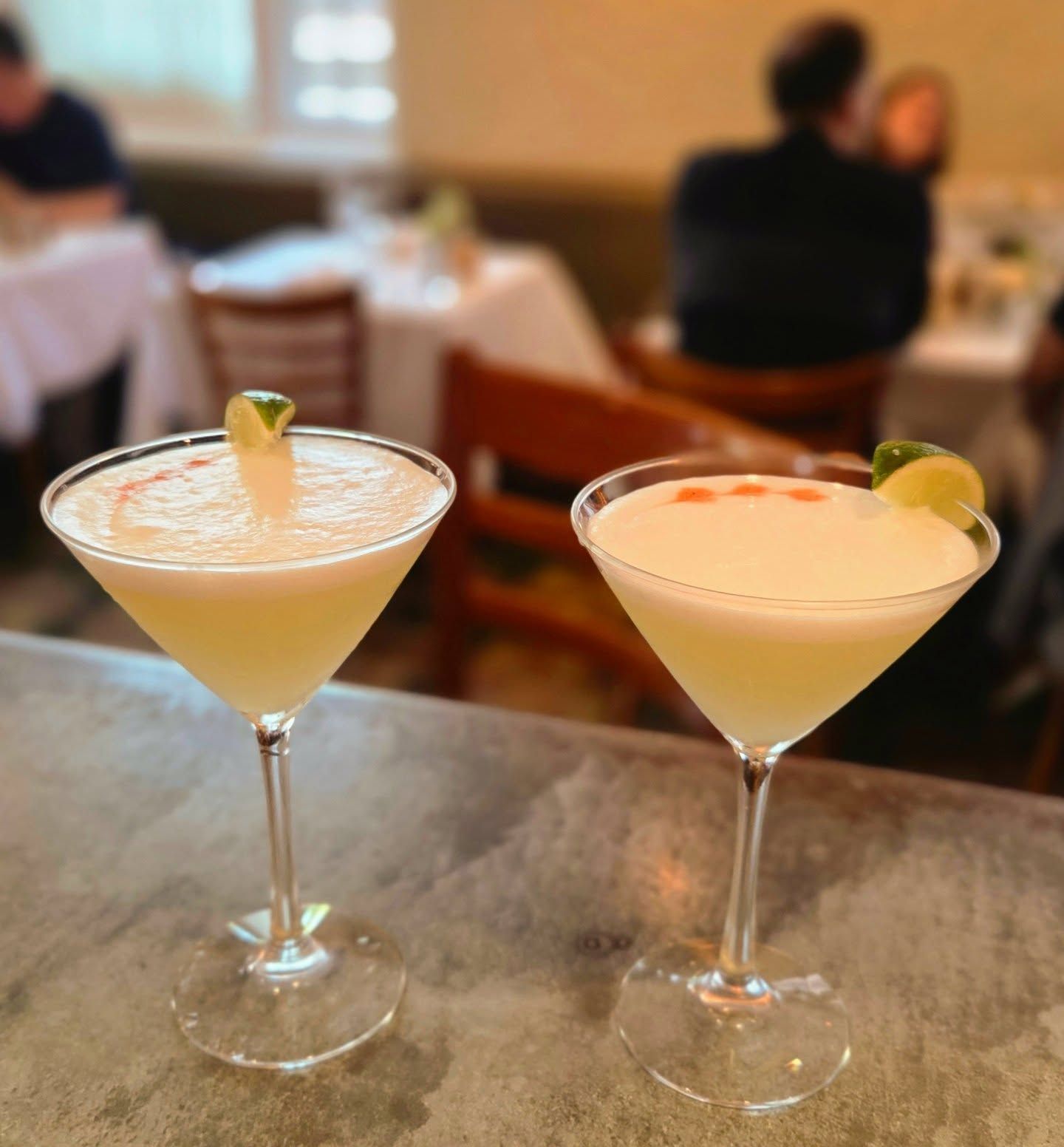 Two martini glasses with pale cocktails and lime garnishes on a bar counter in a restaurant
