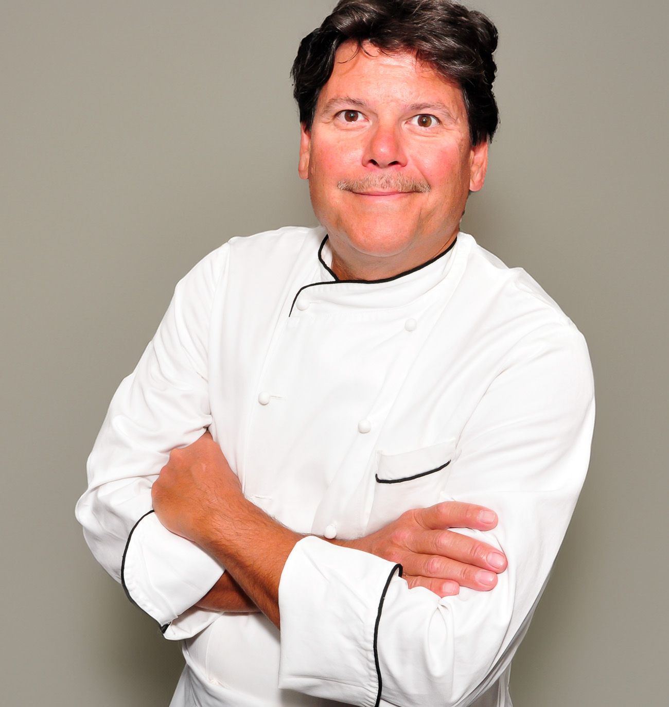 Chef in a white jacket with arms crossed, smiling against a plain gray background