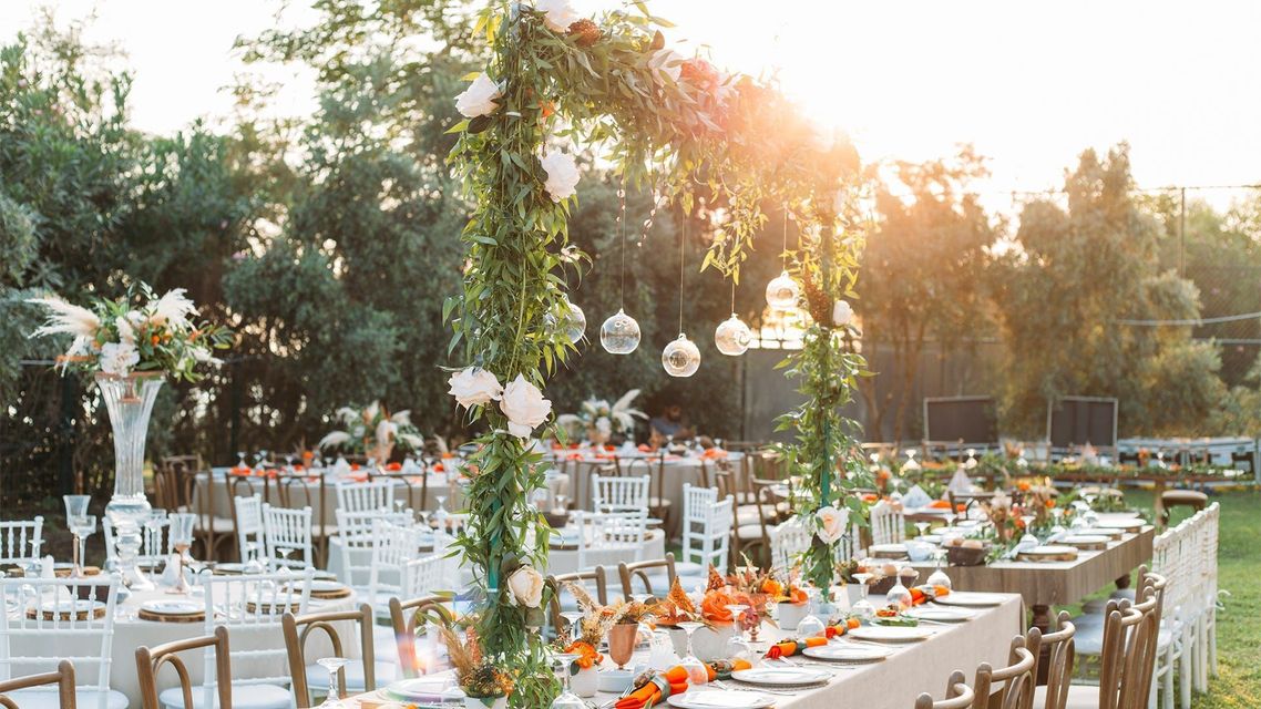 Sunlit outdoor wedding reception with white chairs, long tables, and hanging lanterns among trees