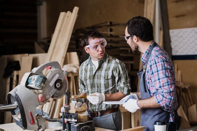 Two people in plaid shirts discuss woodworking in a workshop beside a miter saw.