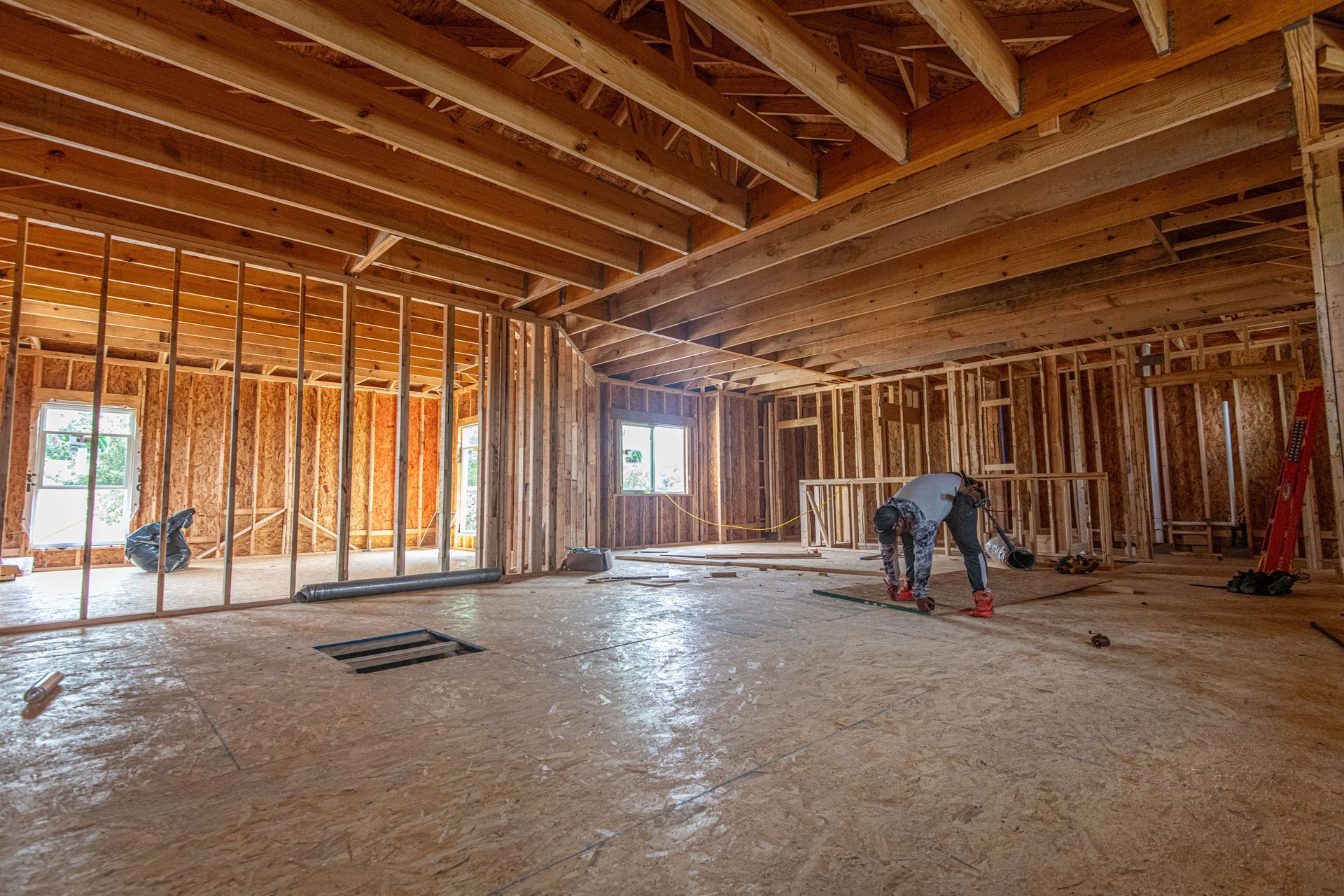 Unfinished house interior with exposed wood framing and a worker crouched on the floor.