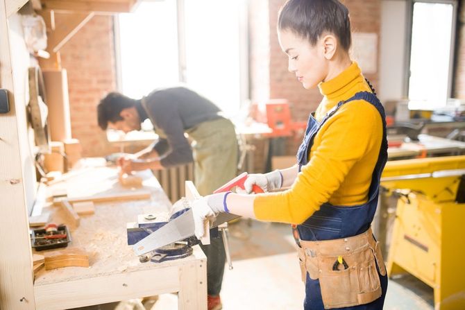 Person using a circular saw in a bright wood workshop, with another worker sanding in the background.