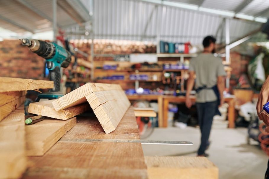 Woodworking shop with stacked lumber, a drill on a bench, and a person walking in the background