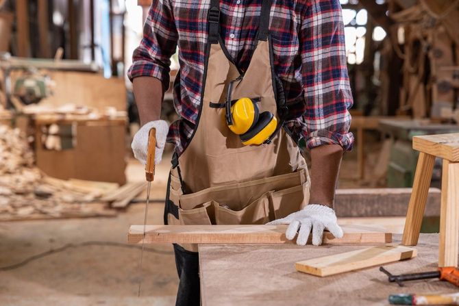 Woodworker in plaid shirt and apron measuring a board in a workshop, with gloves and ear protectors.