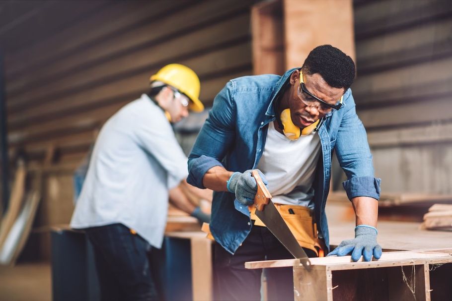 Two workers in a workshop cutting wood at a table, one using a hand saw and wearing gloves and a yellow hard hat.