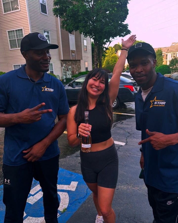 Three people posing outdoors on a wet street, smiling and holding drinks, wearing navy shirts.