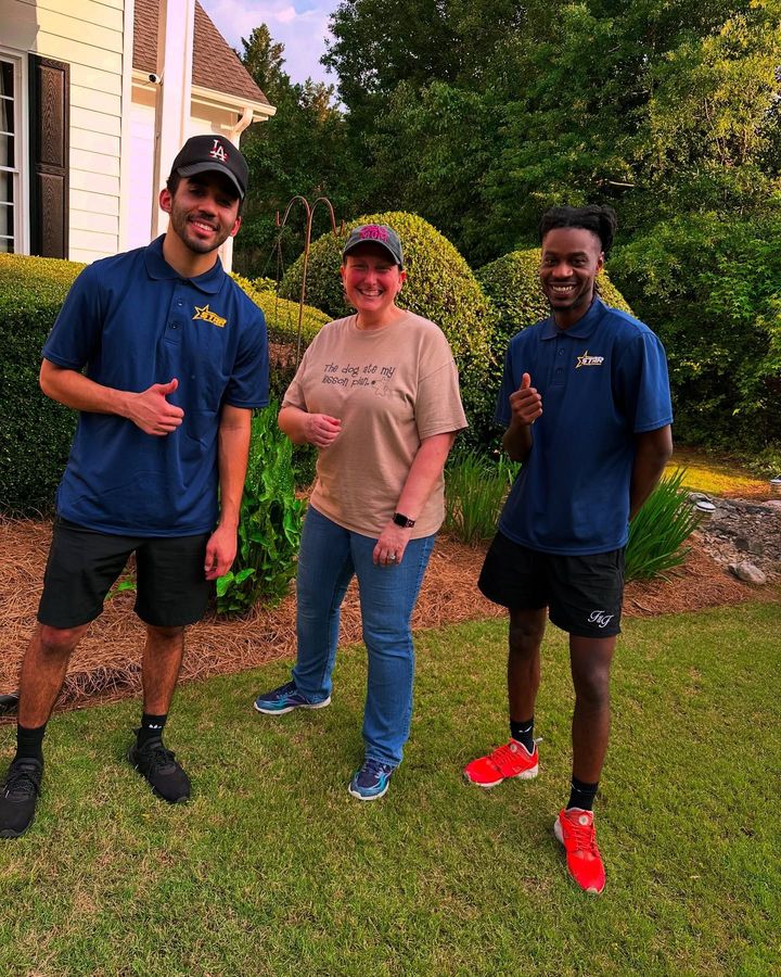 Three people smiling outdoors on a lawn, wearing blue shirts and shorts, posing in front of a house.
