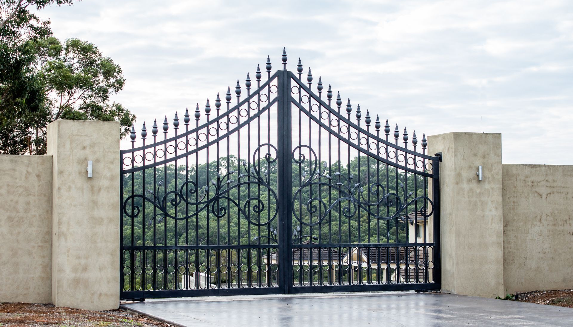 Ornate wrought-iron arched driveway gate with scrollwork and stucco pillars.