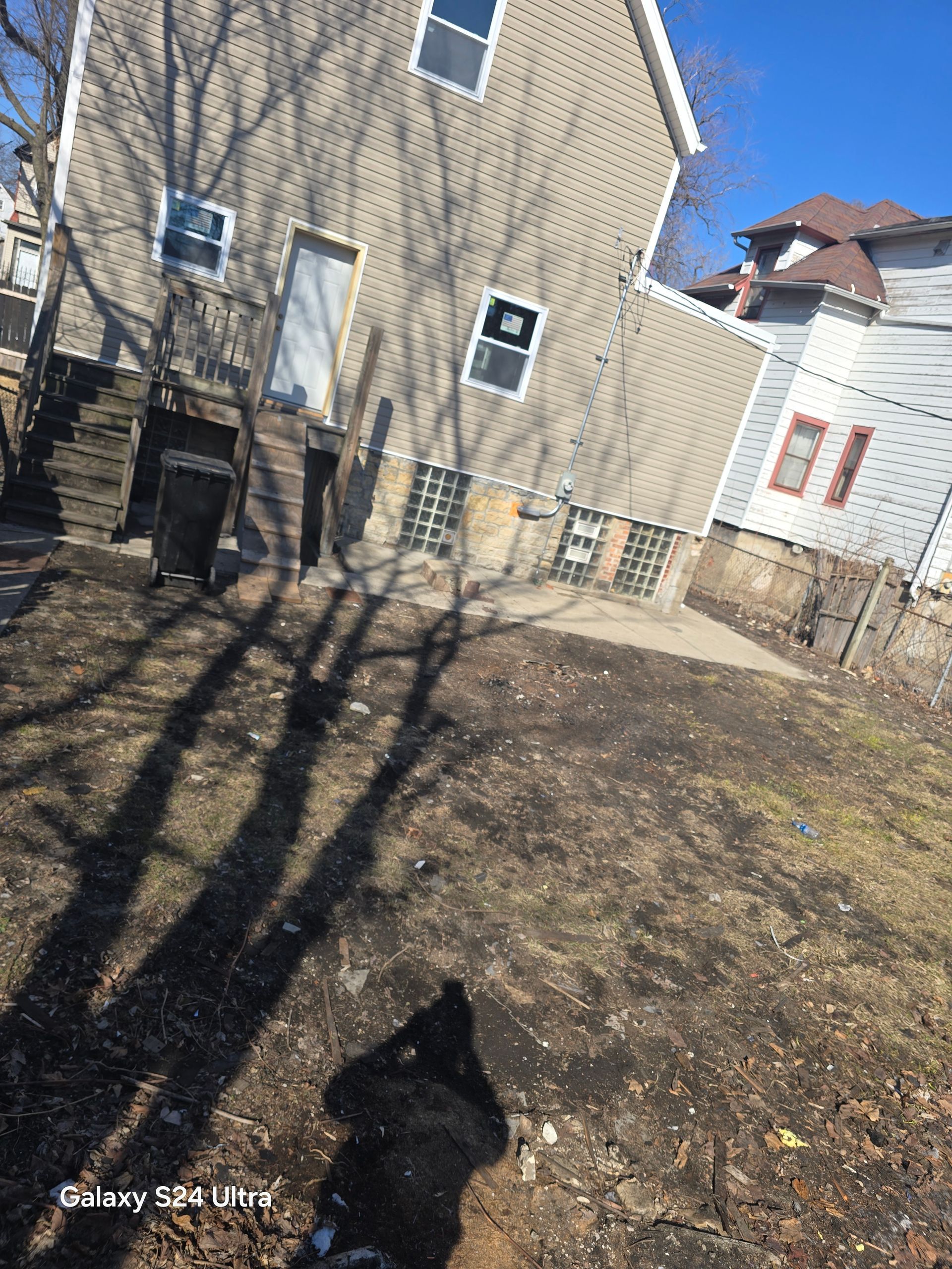 A beige house with a concrete patio and a dirt backyard, with a black dog sitting in the foreground.