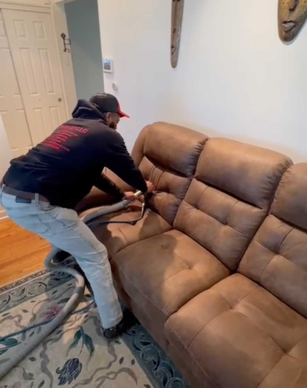 A professional technician uses a vacuum tool to clean a brown fabric sofa in a home setting.