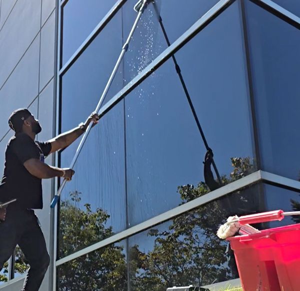 Person washing large windows of a building with long-handled tools, sunny day.