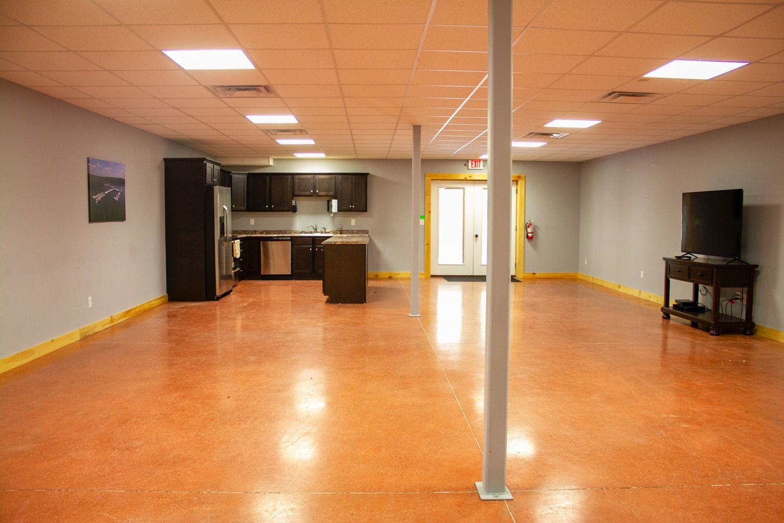 Open space with kitchen, TV, and orange epoxy floor; two support beams, and door.