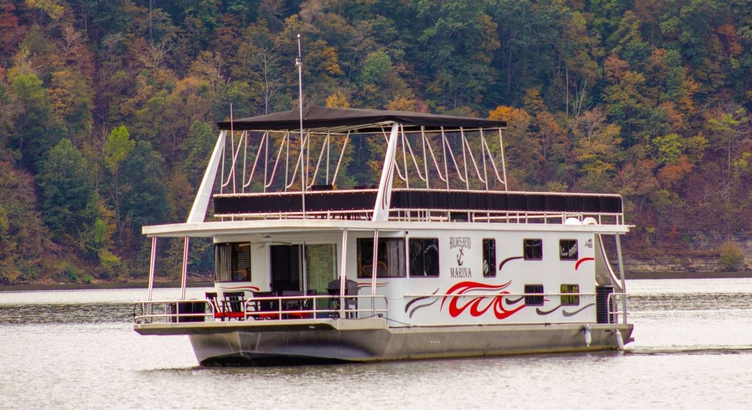 Houseboat on water with red and white decorative flames, trees in the background.