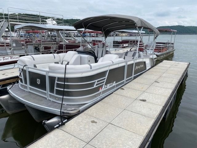 A gray pontoon boat docked at a pier with a canopy. Other boats and the water are in the background.