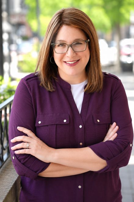 Smiling person in glasses, wearing a purple shirt, standing outdoors with arms crossed.