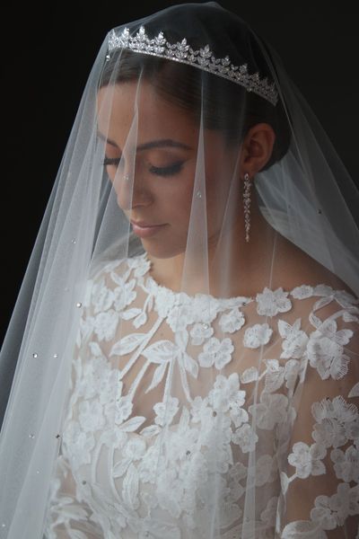 Bride in a white lace dress and veil looking down, softly lit against a dark background