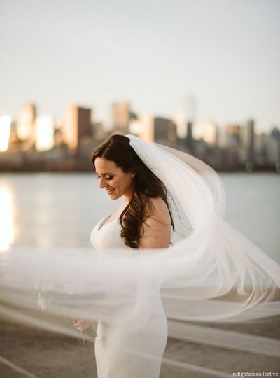 Bride in a white dress and veil standing by the water with a city skyline in the background