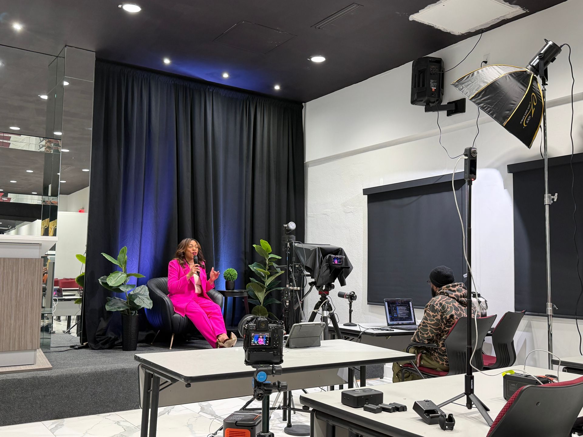 TV studio set with a seated guest in a bright pink outfit, cameras and lighting equipment around her.