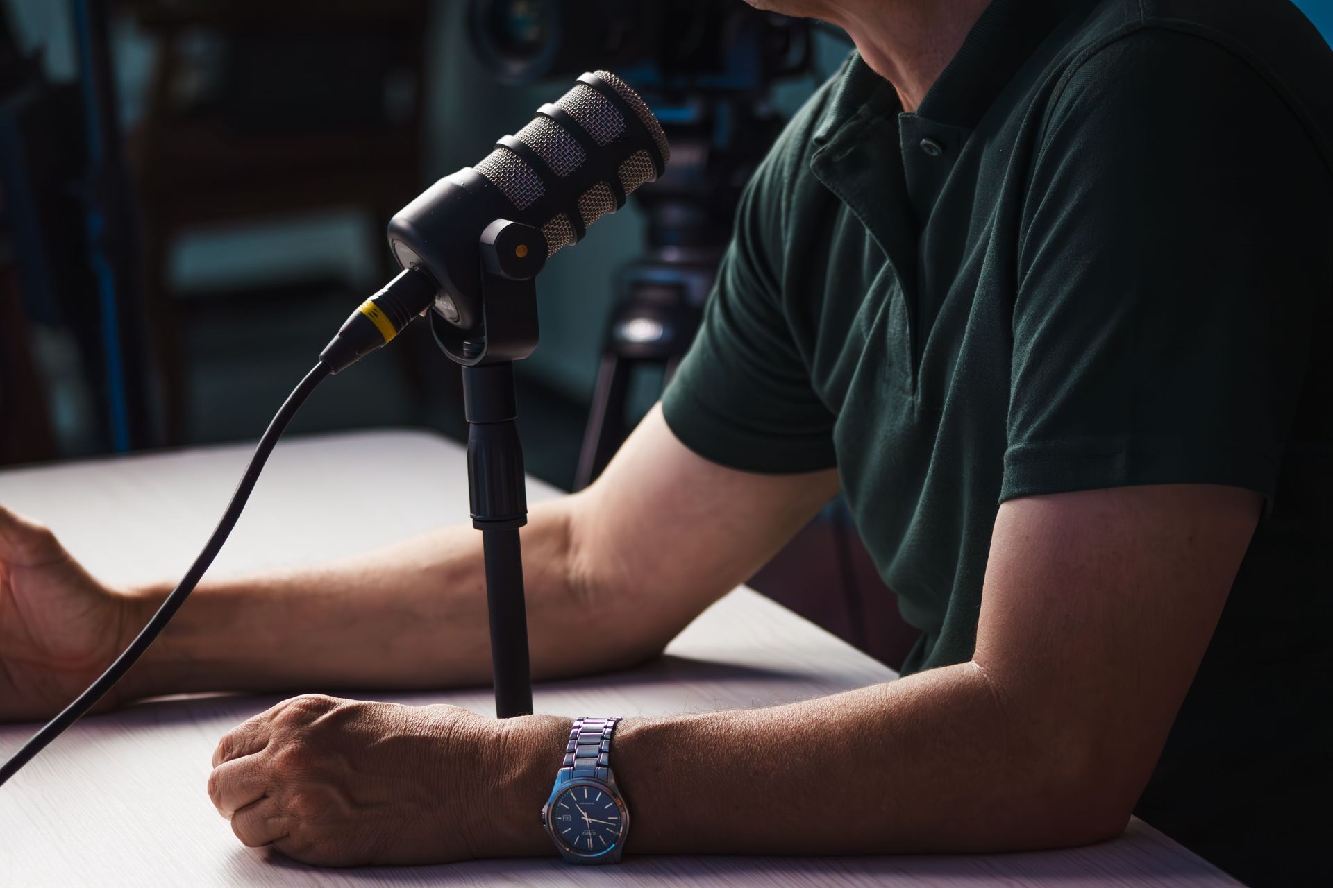 Person speaking into a microphone at a desk, wearing a green shirt and blue watch, in a dim room