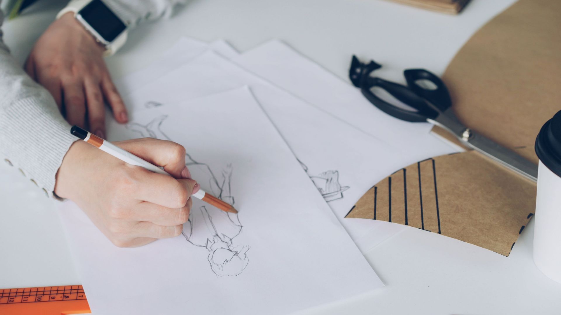 Hands sketching a fashion design on white paper with scissors and cardboard on a desk