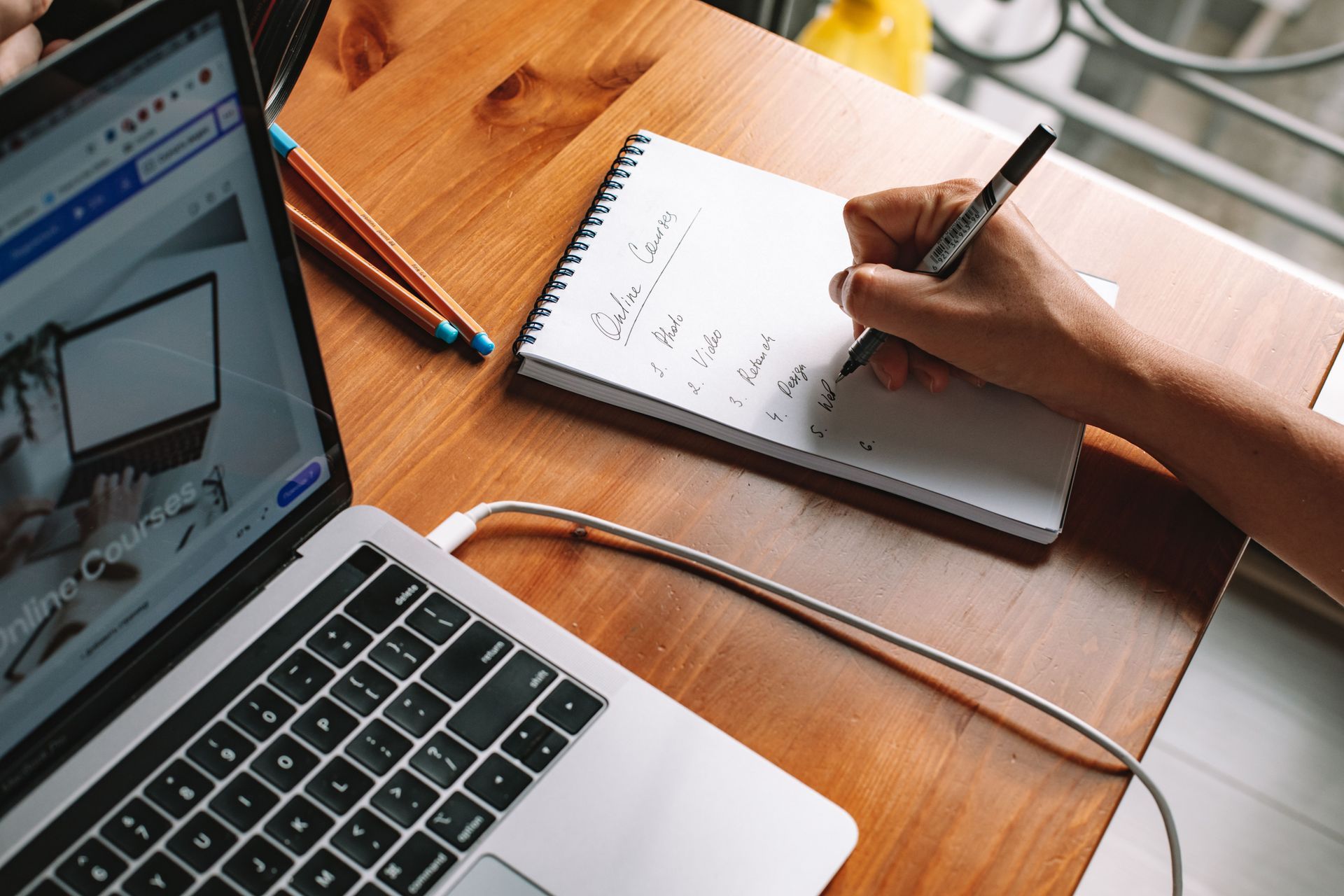 Person writing in a notebook beside a laptop on a wooden desk