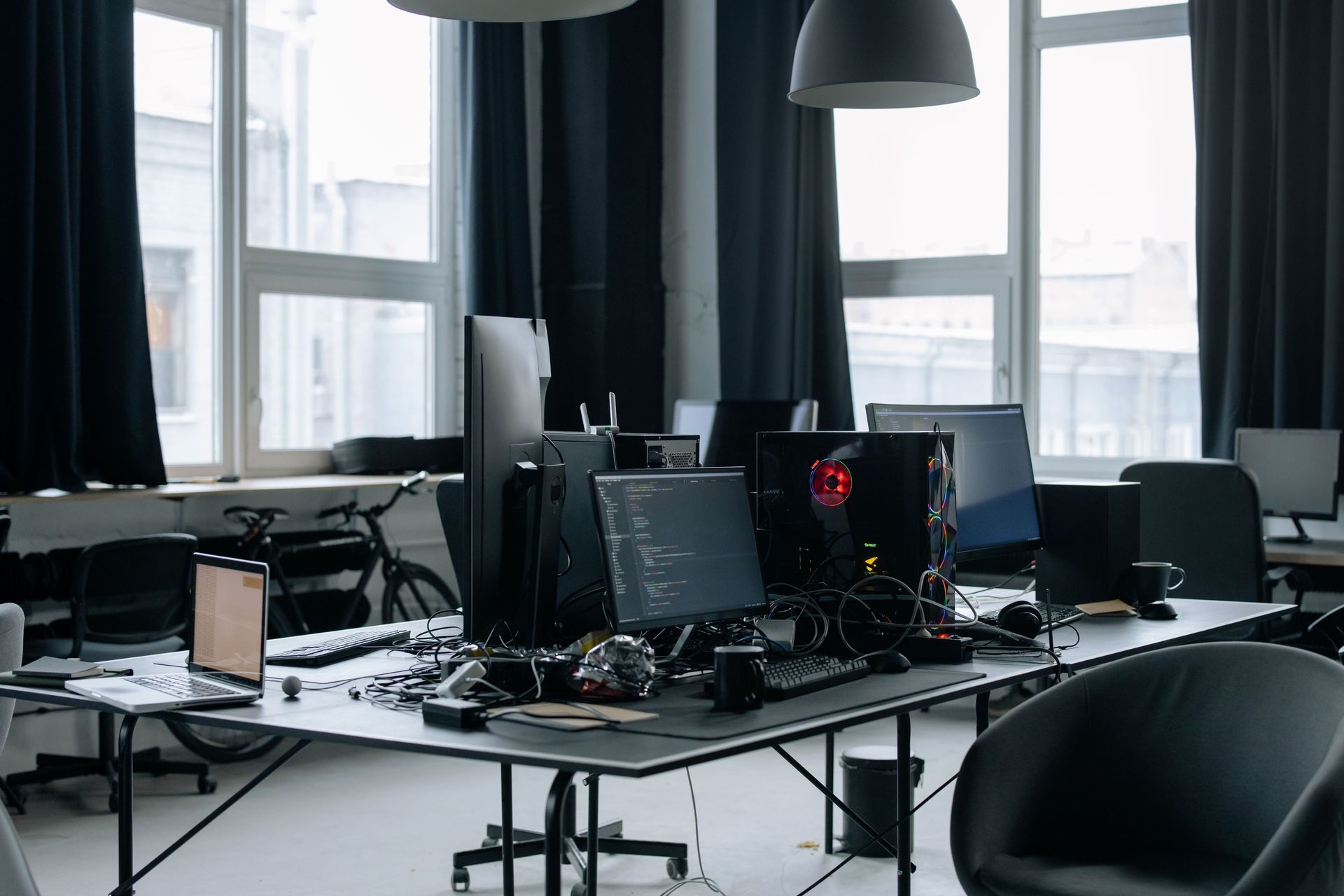 Modern office workspace with multiple computer monitors, desks, cables, and large windows with dark curtains