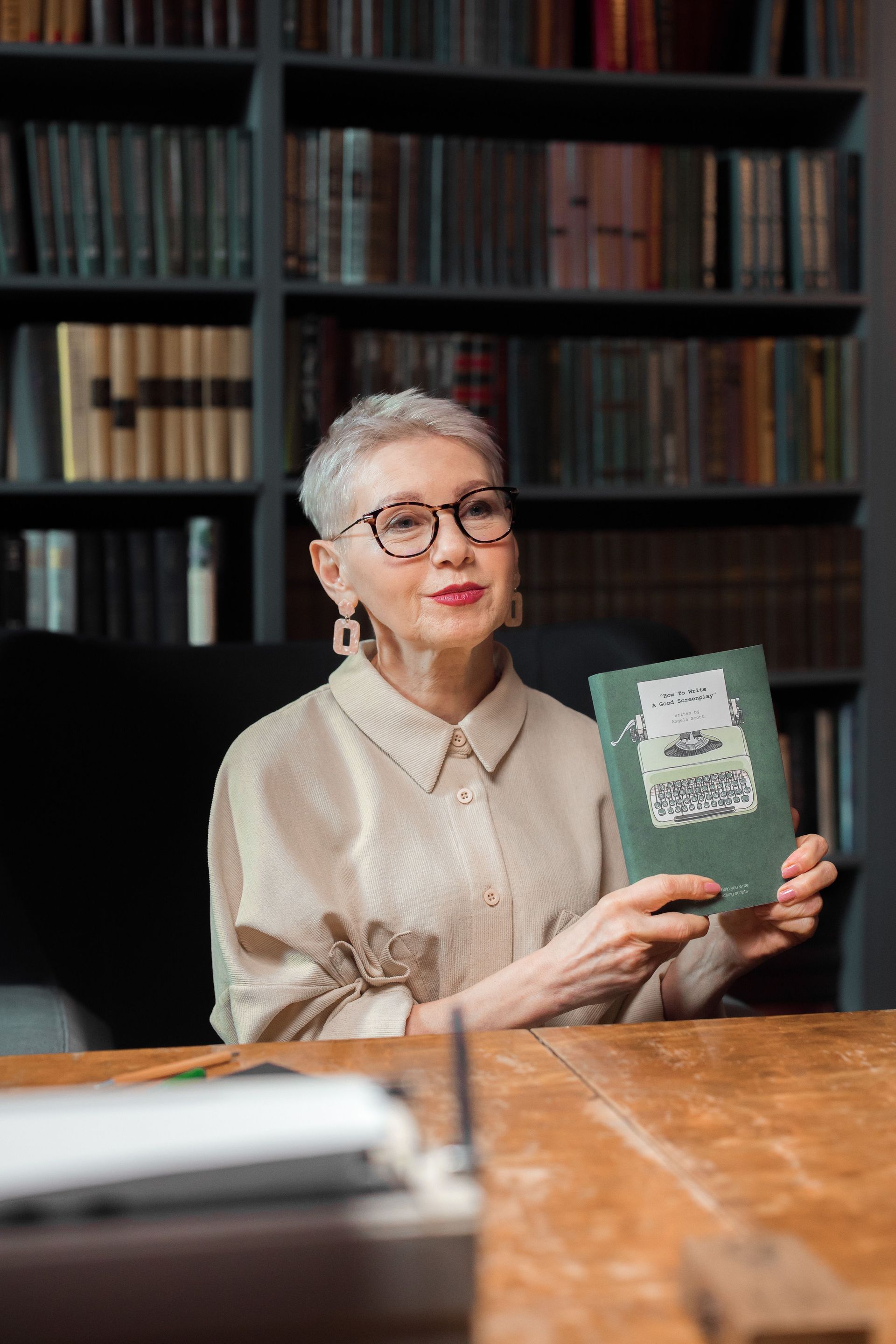 Woman seated in a library holding up a green book beside a wooden table