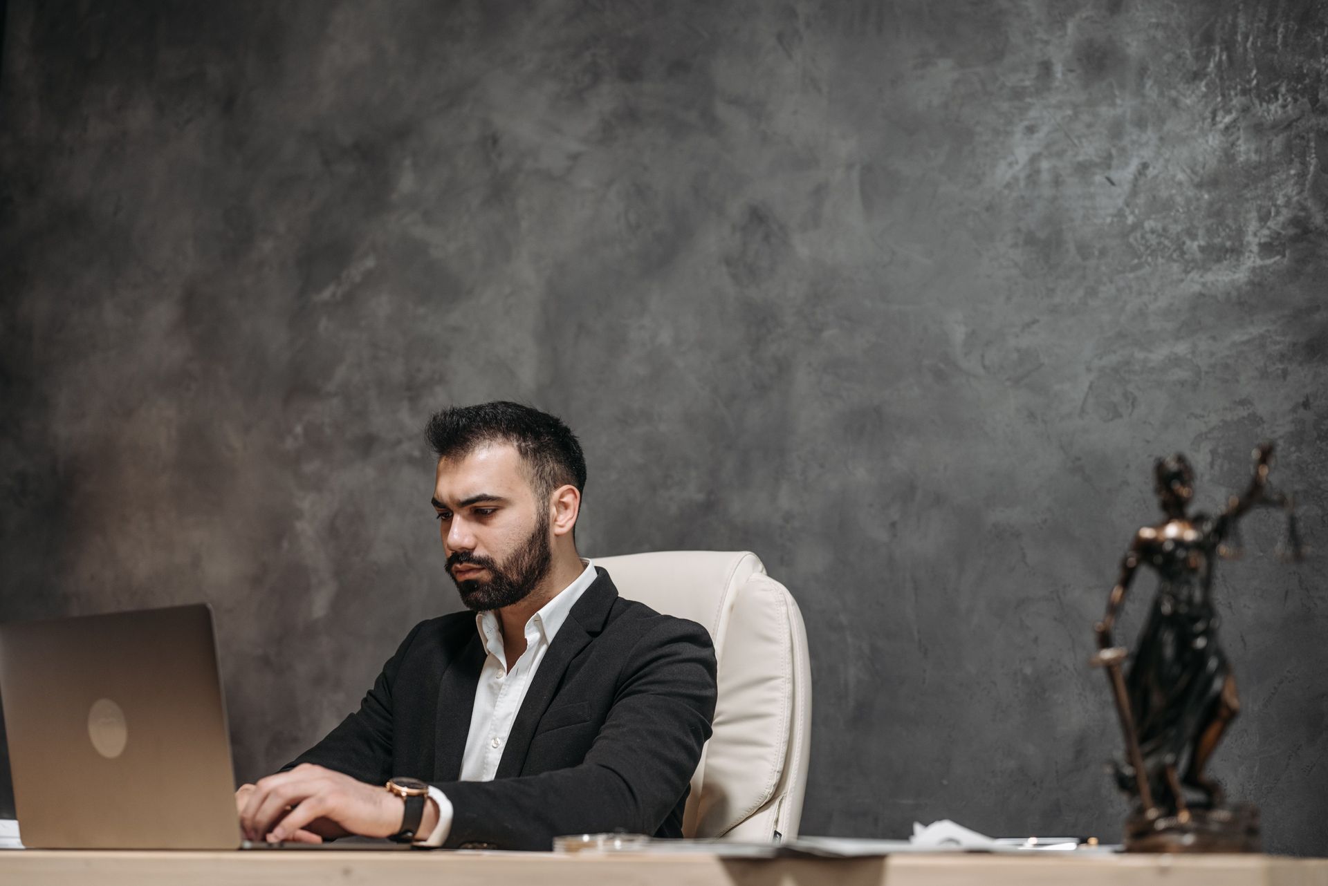 Man in a dark suit working at a laptop in a minimalist office with a dark textured wall