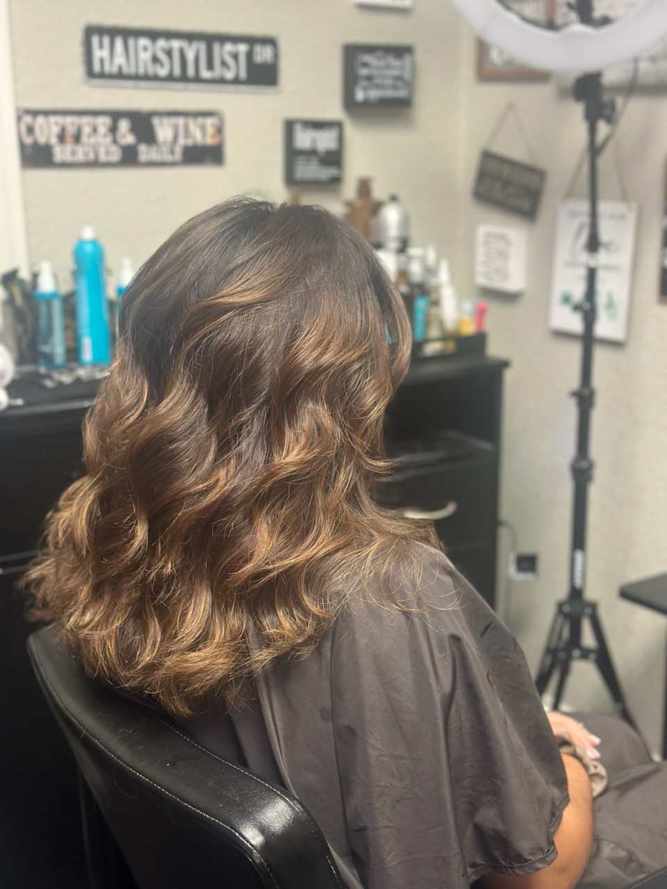 Woman with wavy brown hair sitting in a salon chair, viewed from behind.