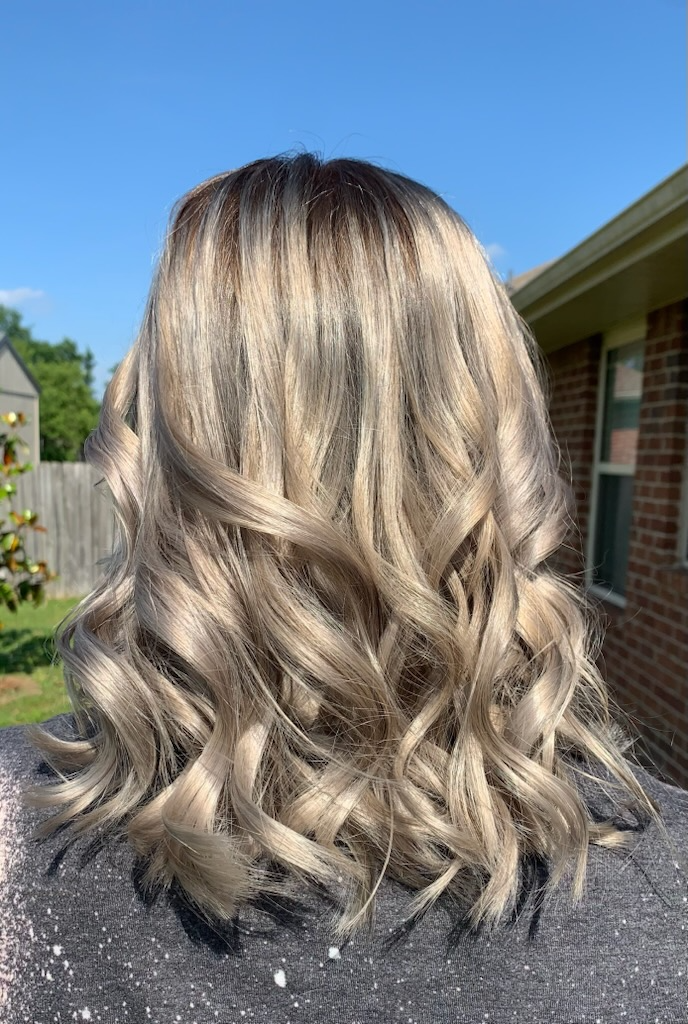 Wavy blonde hair styled in loose curls against a blue sky and brick house backdrop
