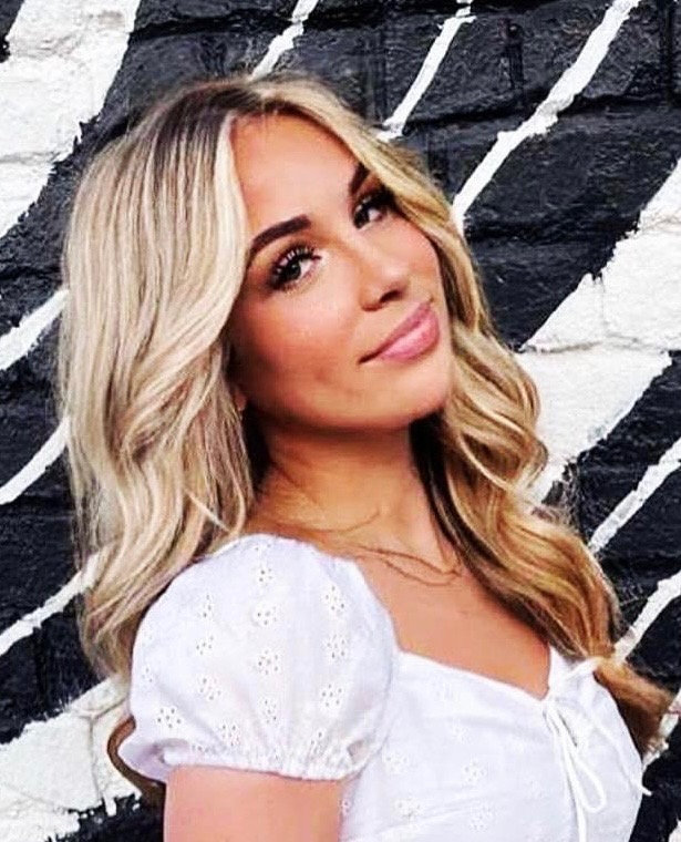 Woman with blonde wavy hair in a white blouse, posing against a black-and-white mural backdrop