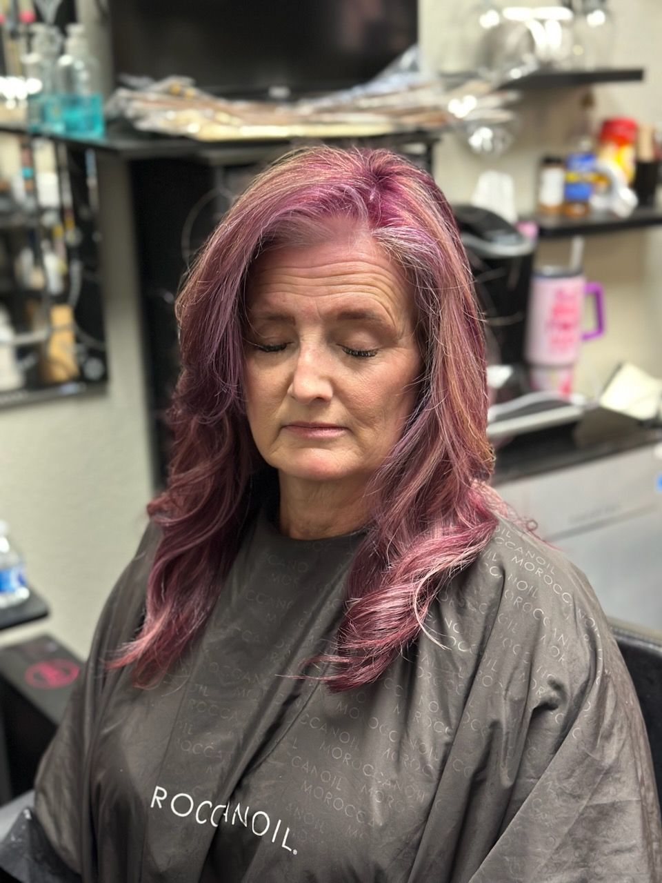 Woman with magenta hair and dark salon cape sitting in a beauty salon chair with eyes closed
