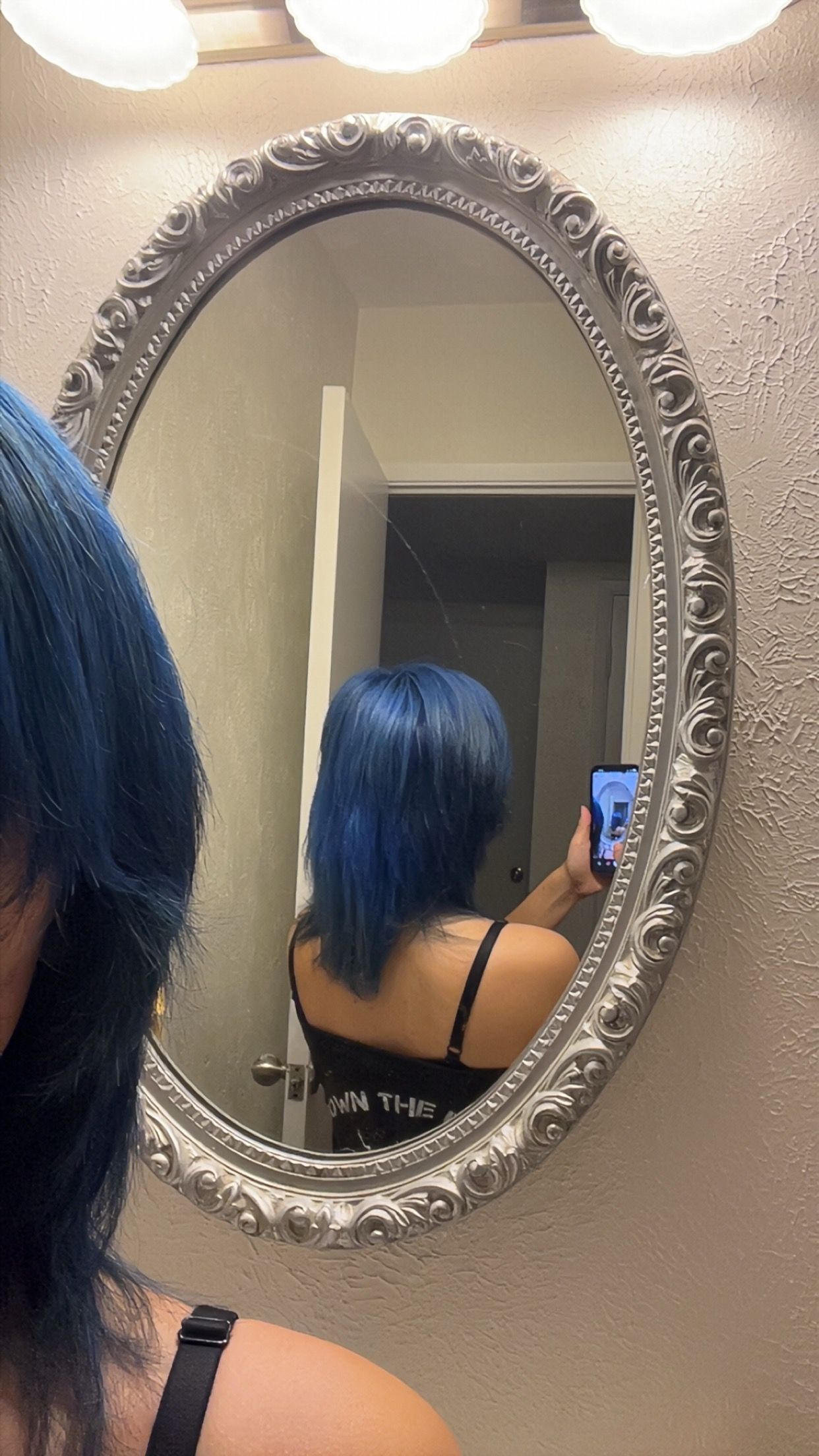 Person with blue hair taking a mirror selfie in a bathroom with an ornate oval mirror.