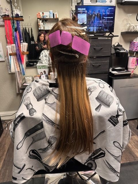 Hair salon client with long brown hair clipped with pink rollers, wearing a salon cape in front of a mirror and TV