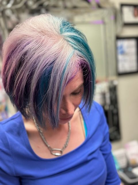 Person with short pastel pink, blue, and purple hair looking down in a salon chair