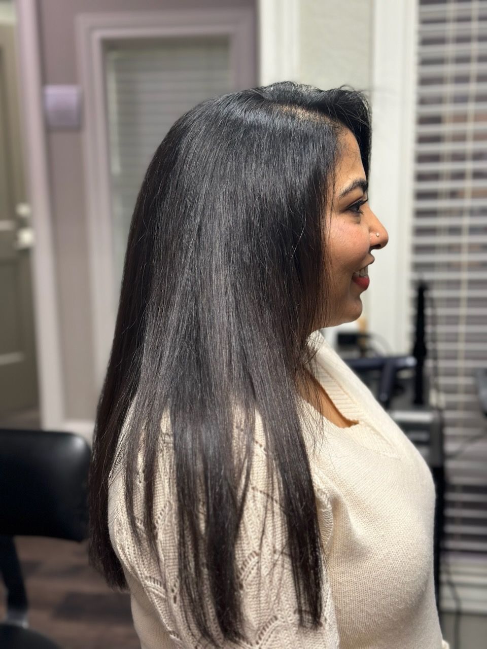 Side view of a woman with long dark hair wearing a white sweater indoors, smiling near a window blinds.