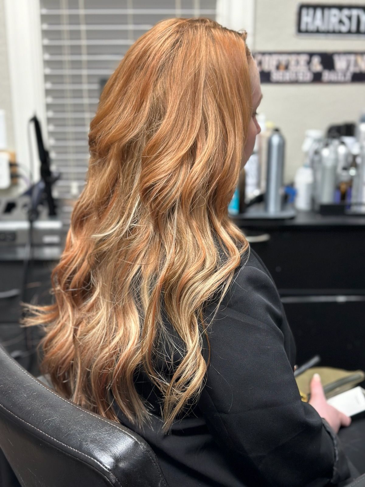 Woman with wavy copper hair seated in a salon chair, viewed from behind.